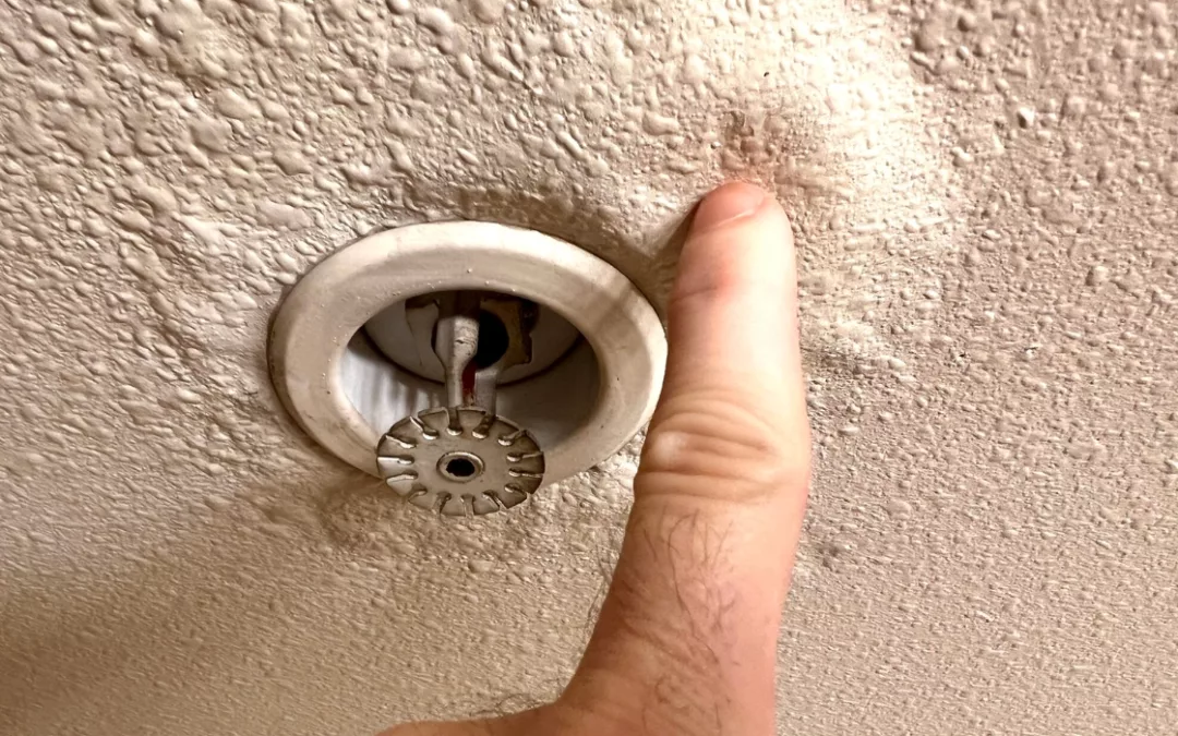 A person touching the ceiling paint that has bubbled up near a water sprinkler to indicate it has a pipe leak